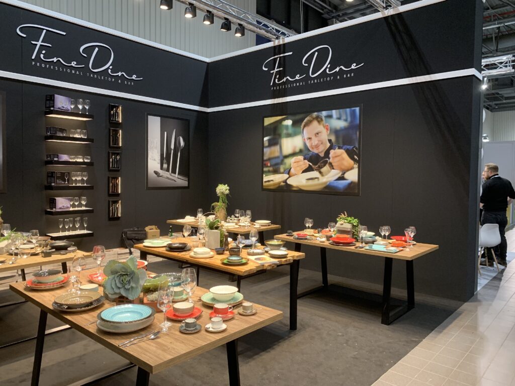 W salonie wystawowym stoją stoły nakryte kolorową zastawą, filiżankami i szklankami: dla wnikliwego obserwatora jest to wyjątkowa ekspozycja porcelany Fine Dine na stołach — kolorowe zestawy pięknie ułożone tak, by wywrzeć jak największe wrażenie. Wzdłuż tylnej ściany ustawiono półki z przyborami kuchennymi, a na czarnej ścianie z logo Fine Dine wisi zdjęcie szefa kuchni serwującego danie. Brutalistyczne. projekt i nadzór Tatjana Kobuszewska