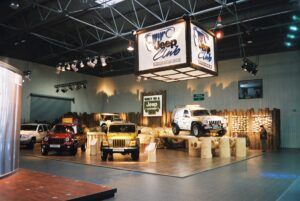 Stoisko targowe Jeep prezentuje wiele pojazdów Jeep na platformach i kłodach, z dużym podświetlanym napisem Jeep Club Poland powyżej i napisem Only in a Jeep na tylnej ścianie. Projekt i nadzór Tatjana Kobuszewska trksystem.com. 