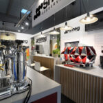 Nowoczesne stoisko Smeg na targach EuroGastro prezentuje błyszczące ekspresy do kawy, w tym chromowany ekspres do kawy i geometryczny czerwony model na ladzie. Stylowe, wyłożone kafelkami stoisko może pochwalić się brandingiem "Smeg Foodservice", lampami wiszącymi i telewizorem powyżej. projekt i nadzór Tatjana Kobuszewska trksystem.com