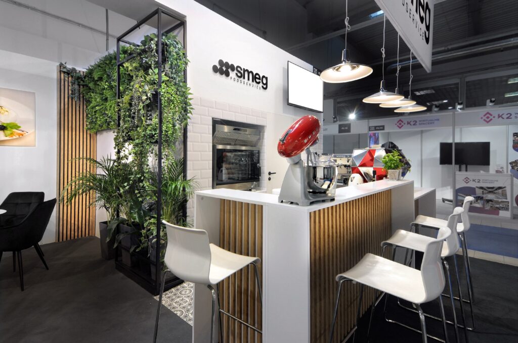 Nowoczesne stoisko wystawowe na targach Ptak Warsaw Expo prezentuje sprzęt AGD marki Smeg, a w jego skład wchodzą: blat z stołkami barowymi, mikser, piekarnik, wiszące lampy, bujne zielone rośliny oraz konstrukcja z drewnianych listew – wszystko to na tle tętniącej życiem atmosfery targów. projekt i nadzór Tatjana Kobuszewska