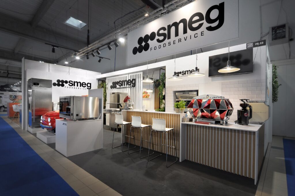 Nowoczesne stoisko wystawiennicze Smeg Foodservice na targach EuroGastro, z białym i drewnianym wystrojem, stołkami barowymi, ekspresami do kawy i kolorowym mozaikowym piecem do pizzy. Logo Smeg wyróżnia się w tej dobrze oświetlonej, zachęcającej przestrzeni. projekt i nadzór pracownia Tatjany Kobuszewskiej