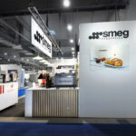 Nowoczesne stoisko SMEG Foodservice na targach Smeg na targach EuroGastro obejmuje ekspresy do kawy, dużą ścianę z logo SMEG, zdjęcie filiżanki kawy i rogalika, akcenty zieleni, eleganckie wyświetlacze i jasne oświetlenie w przestronnej hali. projekt i nadzór Tatjana Kobuszewska trksystem.com