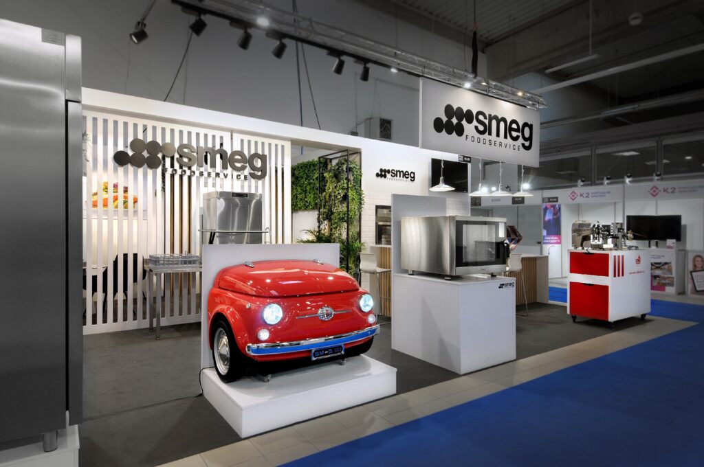 Nowoczesne stoisko targowe Smeg na targach EuroGastro prezentuje urządzenia Smeg, w tym czerwoną lodówkę retro w kształcie zabytkowego samochodu, piekarniki i sprzęt kuchenny. Markowe znaki Smeg wiszą nad czystym, otwartym układem z niebieską wykładziną podłogową. projekt i nadzór Tatjana Kobuszewska