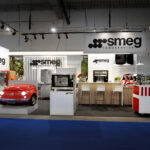 Eleganckie stoisko Smeg na targach EuroGastro prezentuje urządzenia kuchenne, w tym czerwoną lodówkę w kształcie samochodu, lodówkę ze stali nierdzewnej, piekarniki, ekspresy do kawy i nowoczesną drewnianą ladę ze stołkami pod widocznym brandingiem Smeg. projekt i nadzór Tatjana Kobuszewska trksystem.com