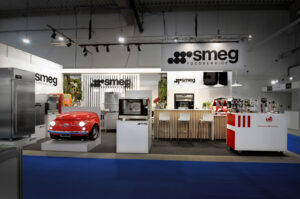 Eleganckie stoisko Smeg na targach EuroGastro prezentuje urządzenia kuchenne, w tym czerwoną lodówkę w kształcie samochodu, lodówkę ze stali nierdzewnej, piekarniki, ekspresy do kawy i nowoczesną drewnianą ladę ze stołkami pod widocznym brandingiem Smeg. projekt i nadzór Tatjana Kobuszewska trksystem.com