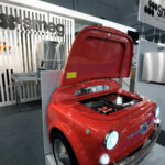 Czerwona lodówka SMEG w stylu retro, zaprojektowana tak, aby przypominała przód zabytkowego Fiata 500, z reflektorami i otwartą maską, jest prezentowana na stoisku targowym z brandingiem Smeg na targach EuroGastro i urządzeniami kuchennymi ze stali nierdzewnej w tle. projekt i nadzór Tatjana Kobuszewska trksystem.com