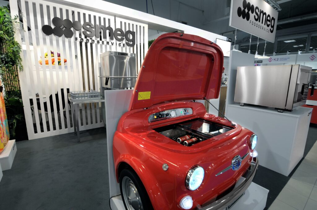 Czerwone urządzenie kuchenne SMEG w kształcie przodu klasycznego samochodu fiat 500 jest prezentowane z otwartą maską, odsłaniając elementy kuchenne wewnątrz. W tle na targach EuroGastro widoczne są białe panele wystawowe i urządzenia ze stali nierdzewnej. projekt i nadzór Tatjana Kobuszewska