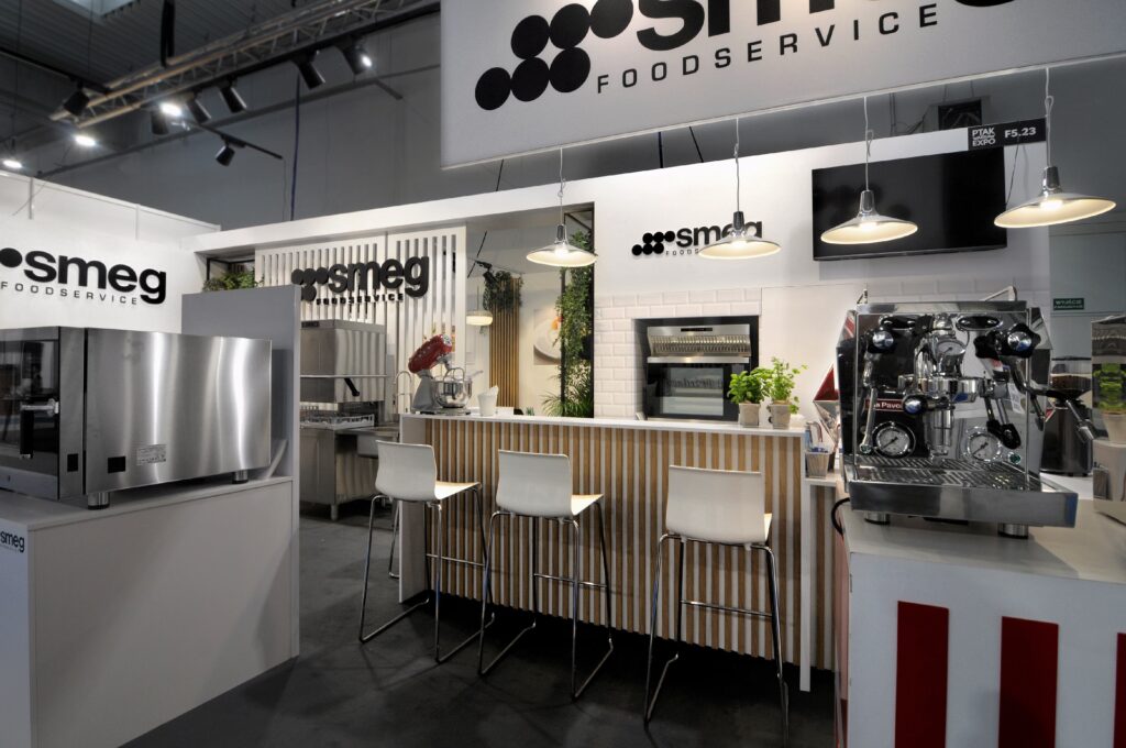 Nowoczesne stoisko gastronomiczne SMEG na targach EuroGastro, wyposażone w urządzenia kuchenne ze stali nierdzewnej, ladę z trzema białymi stołkami barowymi, lampy wiszące powyżej i branding SMEG. Ekspres do kawy i piekarnik są wyraźnie wyeksponowane. projekt i nadzór Tatjana Kobuszewska