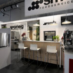 Nowoczesne stoisko w stylu kawiarnianym na targach Smeg na targach EuroGastro obejmuje urządzenia gastronomiczne SMEG, białe stołki barowe przy ladzie w paski, ekspres do kawy, piekarniki i wiszące lampy w jasnej, minimalistycznej przestrzeni z widocznym brandingiem SMEG powyżej. projekt i nadzór Tatjana Kobuszewska trksystem.com