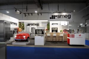 Nowoczesne stoisko wystawowe firmy Smeg Foodservice, zaprojektowane w ramach projektu i nadzoru Tatjana Kobuszewska trksystem.com, oferuje urządzenia ze stali nierdzewnej, ladę w stylu retro w kolorze czerwonym, drewniane hokery barowe i ekspresy do kawy w dobrze oświetlonej przestrzeni wewnętrznej. projekt i nadzór Tatjana Kobuszewska trksystem.com