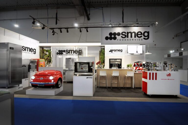 Nowoczesne stoisko wystawowe firmy Smeg Foodservice, zaprojektowane w ramach projektu i nadzoru Tatjana Kobuszewska trksystem.com, oferuje urządzenia ze stali nierdzewnej, ladę w stylu retro w kolorze czerwonym, drewniane hokery barowe i ekspresy do kawy w dobrze oświetlonej przestrzeni wewnętrznej. projekt i nadzór Tatjana Kobuszewska trksystem.com