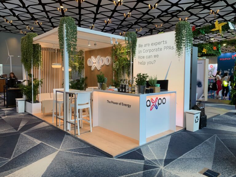 Stoisko konferencyjne Axpo ma białe ściany, drewniane akcenty, wiszące rośliny i logo Axpo. To zachęcające /stoiska-konferencyjne/ obejmuje ladę ze stołkami, miejsce do siedzenia i znaki podkreślające wiedzę specjalistyczną w zakresie korporacyjnych umów PPA i rozwiązań energetycznych. projekt i nadzór Tatjana Kobuszewska trksystem.com