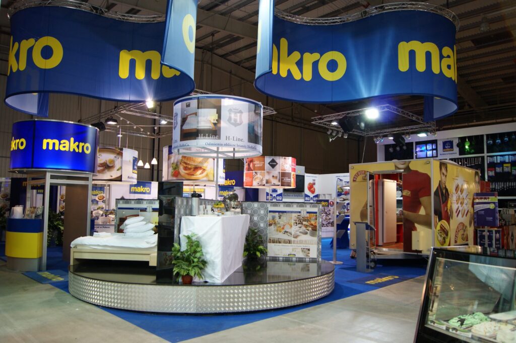 Stoisko targowe z dużymi niebieskimi i żółtymi szyldami „makro”, stoiska z produktami kuchennymi i żywnością uliczną, materiały promocyjne, rośliny doniczkowe i różne zdjęcia związane z jedzeniem w tle. projekt i nadzór Tatjana Kobuszewska trksystem.com