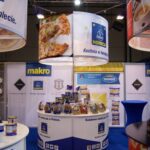 Stoisko targowe z marką Makro i Horeca prezentuje różne pakowane produkty spożywcze, w tym opcje jedzenia ulicznego. Duże plakaty pokazują pizzę i inne potrawy. Stoisko ma niebieskie wykładziny i lady z produktami i materiałami promocyjnymi. projekt i nadzór Tatjana Kobuszewska trksystem.com