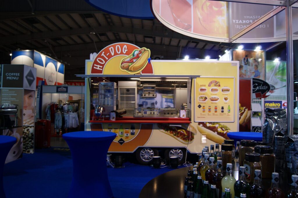 Jasno oświetlony uliczny food hot dog truck z dużym ilustrowanym znakiem hot dog jest zaparkowany w pomieszczeniu na imprezie. Niebieskie stoliki koktajlowe i butelki są na pierwszym planie; w tle widać ludzi i różne stragany. projekt i nadzór Tatjana Kobuszewska trksystem.com