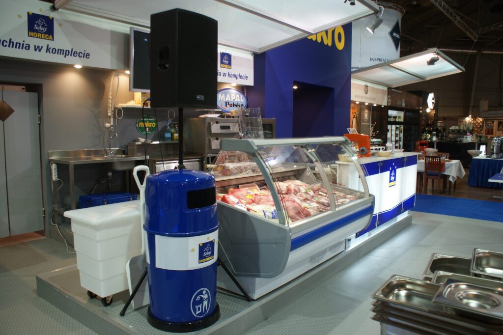 Na targach EuroGastro w pomieszczeniach ustawiono chłodnię mięsną wypełnioną różnymi kawałkami mięsa. W pobliżu znajduje się niebieski kosz na śmieci, duży głośnik i zlew ze stali nierdzewnej, wszystko pod niebiesko-białym szyldem. projekt i nadzór Tatjana Kobuszewska trksystem.com