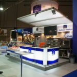 Nowoczesne stoisko targowe EuroGastro z niebiesko-białymi ladami, ekspozycjami żywności, wyposażeniem kuchni i szyldami promocyjnymi w języku polskim wyróżnia się wśród innych stoisk w dużej hali wystawienniczej. projekt i nadzór Tatjana Kobuszewska trksystem.com