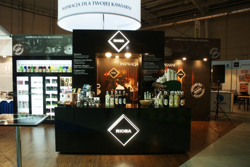 Nowoczesne stoisko z kawą z logo „RIOBA”, podświetlanymi szyldami i różnymi produktami kawowymi; barista pracuje za ladą na targach EuroGastro. projekt i nadzór Tatjana Kobuszewska trksystem.com