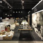 Nowoczesny showroom ze starannie ustawionymi stołami, na których eksponowana jest elegancka zastawa stołowa, szklanki i sztućce. Białe krzesła i skrzynki dodają uroku wystrojowi. Na otaczających czarnych ścianach wiszą duże czarno-białe zdjęcia szefa kuchni. projekt i nadzór Tatjana Kobuszewska trksystem.com