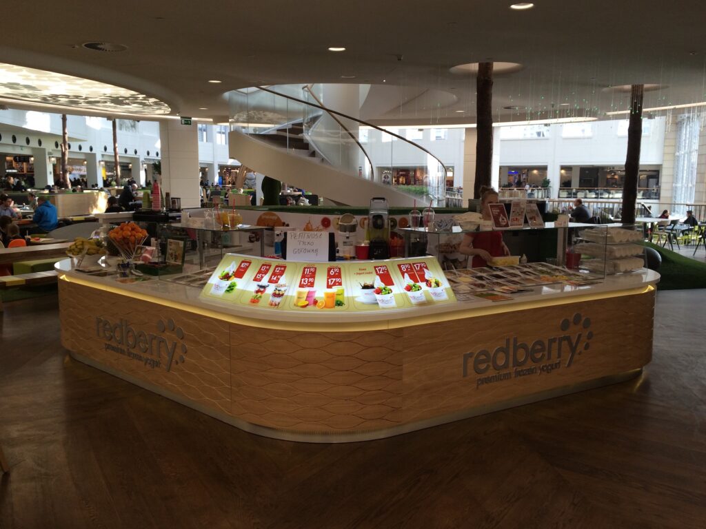 Nowoczesny kiosk z mrożonym jogurtem o nazwie „Redberry”, zaprojektowany jako elegancka wyspa handlowa, przyciąga uwagę jasnymi tablicami z nazwami smaków i dodatków. Kiosk znajduje się w centrum handlowym w pobliżu spiralnych schodów, a ludzie spożywają posiłki i spacerują w delikatnym oświetleniu. projekt i nadzór Tatjana Kobuszewska