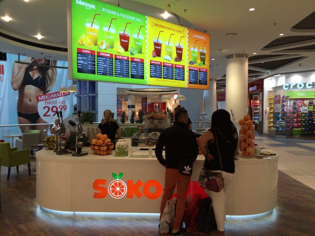 Bar z sokami o nazwie SOKOJU oferuje jasne, kolorowe menu oraz efektowną ekspozycję towaru na wyspie, gdzie pomarańcze są wyeksponowane w szklanych pojemnikach. Dwie osoby składają zamówienie przy ladzie w tym centrum handlowym, a w tle widać inne sklepy. projekt i nadzór Tatjana Kobuszewska
