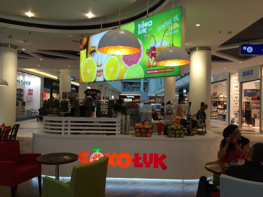 Bar z sokami o nazwie SOKOŁYK, mieszczący się w nowoczesnym centrum handlowym, wyróżnia się eleganckim projektem wyspy handlowej. Na ladzie leżą świeże pomarańcze i jabłka, a na cyfrowym ekranie nad nią wyświetlane są kolorowe zdjęcia owoców i napojów. Ludzie odpoczywają przy pobliskich stolikach, mając w tle sklepy. projekt i nadzór Tatjana Kobuszewska