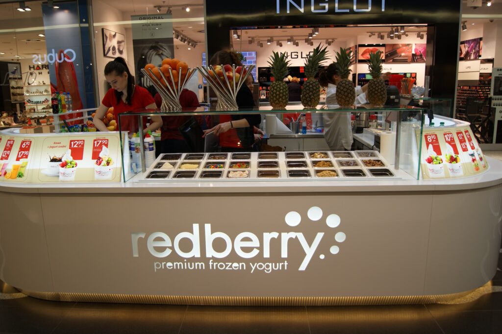 Na ladzie z mrożonymi jogurtami, oznaczonej jako „Redberry Premium Frozen Yogurt”, na jej żywej wyspie handlowej wyeksponowano wiele dodatków, w tym owoce i cukierki. Personel przygotowuje zamówienia, a nad kolorowymi owocami na wyspie handlowej widnieją jasne karty z menu. projekt i nadzór Tatjana Kobuszewska