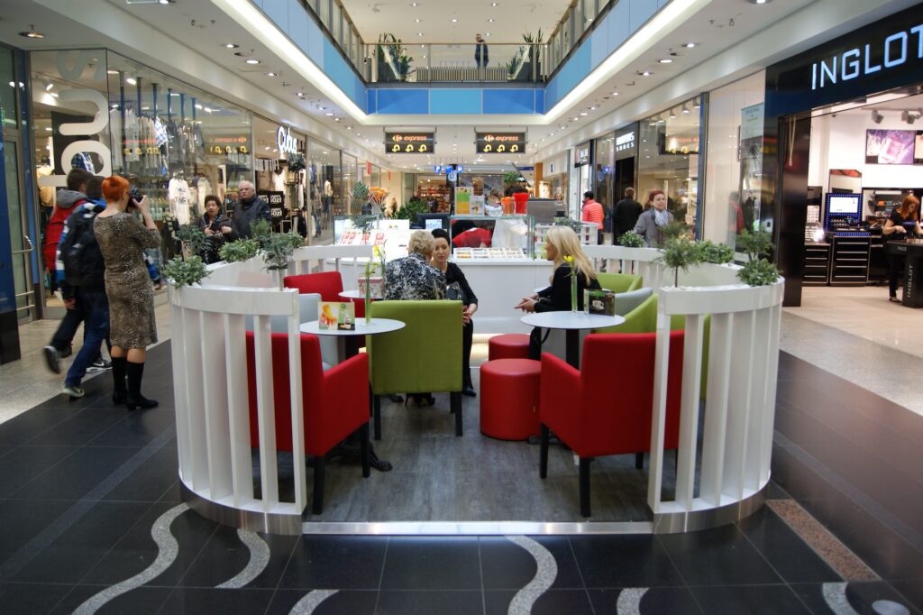 Nowoczesne wnętrze centrum handlowego, w którym ludzie spacerują i robią zakupy. Na środku znajduje się strefa wypoczynkowa „wyspa handlowa” z czerwonymi i zielonymi krzesłami, na których siedzi kilka osób. W tle widać witryny różnych sklepów, w tym Inglot. projekt i nadzór Tatjana Kobuszewska
