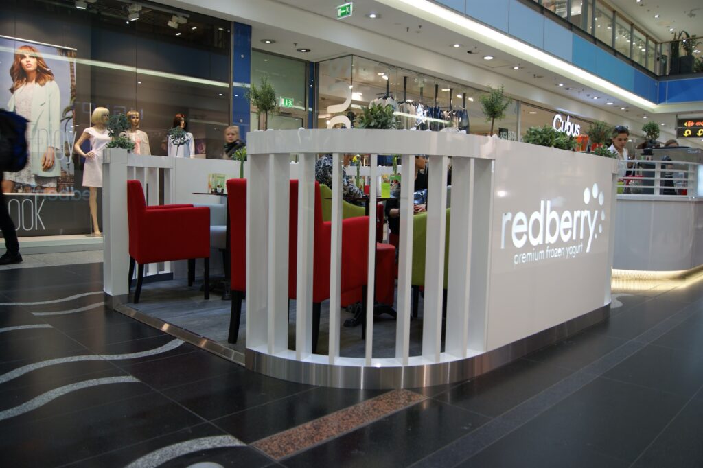 Nowoczesny kiosk typu „wyspa handlowa” z białym ladą z napisem „Redberry Premium Frozen Yogurt”. W środku znajdują się zielone i czerwone krzesła, stoliki oraz rośliny doniczkowe. W pobliżu ludzie przechodzą obok witryn sklepów odzieżowych, w których wystawiono manekiny i reklamy modowe. projekt i nadzór Tatjana Kobuszewska