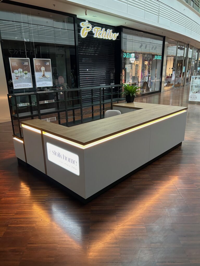 W centrum handlowym, na wypolerowanej drewnianej podłodze, stoi nowoczesna, minimalistyczna wyspa handlowa z białymi panelami i drewnianym blatem, otoczona delikatnym oświetleniem LED. Na blacie stoi zielona roślina, a w tle widać sklepy. projekt i nadzór Tatjana Kobuszewska