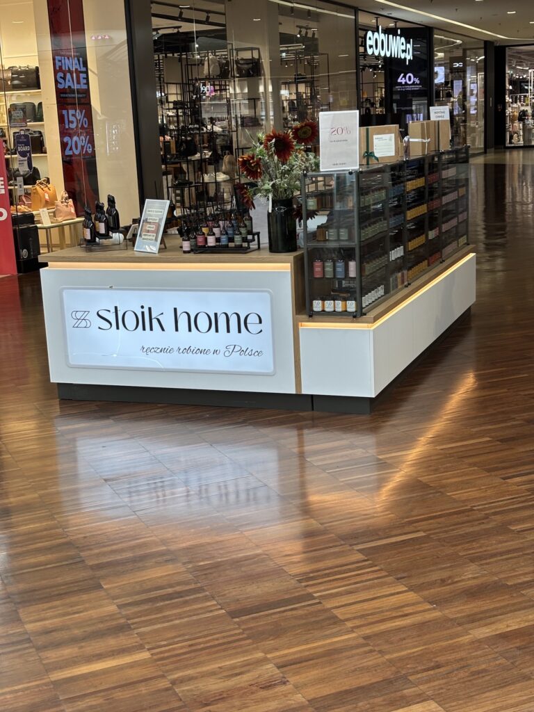 W kiosku w centrum handlowym wystawiono różne produkty do aromatyzowania wnętrz oraz kwiaty, co stanowi atrakcyjną ekspozycję towaru na wyspie. Na szyldzie widnieje napis „Sloik Home – ręcznie robione w Polsce”. W pobliżu widać promocyjne tabliczki z ofertami oraz podłogę o wzorze imitującym ciemne drewno. projekt i nadzór Tatjana Kobuszewska