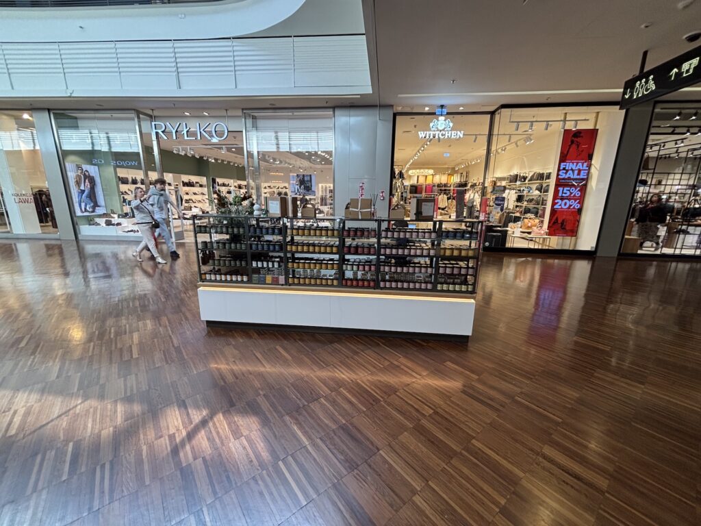 We wnętrzu centrum handlowego znajduje się centralna wyspa handlowa, na której sprzedawane są drobne artykuły. Za nią widoczne są witryny sklepowe: „RYŁKO” z ekspozycją obuwia, „THE BODY SHOP” z napisem „Wyprzedaż końcowa” oraz sklep z odzieżą. Po lewej stronie, po drewnianej podłodze, przechodzą dwie osoby. projekt i nadzór Tatjana Kobuszewska