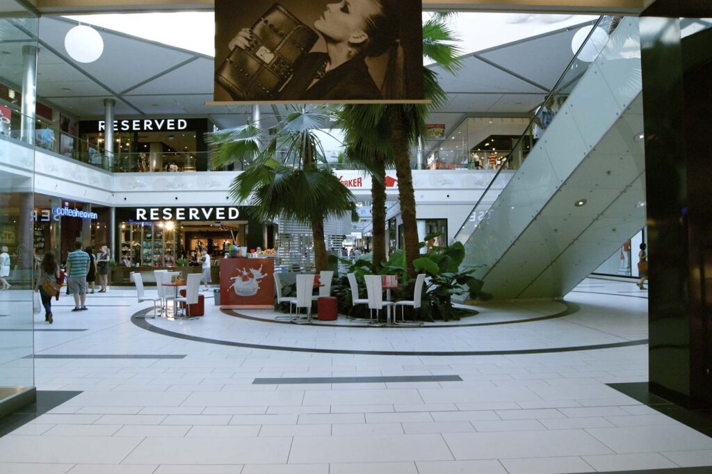 Nowoczesne centrum handlowe posiada szklane balustrady, palmy, białe podłogi wyłożone płytkami, schody ruchome, strefę wypoczynkową ze stolikami i krzesłami, kilka sklepów — w tym dwa sklepy „Reserved” — oraz duży czarno-biały plakat modowy wiszący nad nimi, idealny do każdego projektu wyspy handlowej. projekt i nadzór Tatjana Kobuszewska