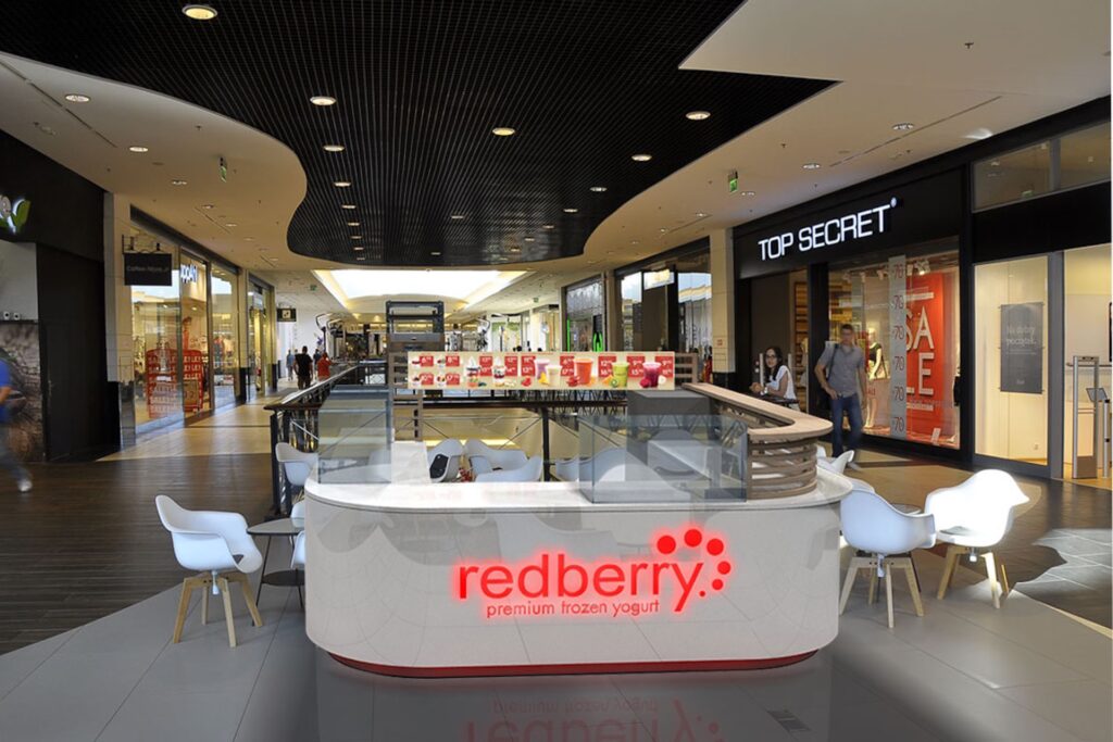 W nowoczesnym centrum handlowym stoi elegancki kiosk typu „wyspa handlowa” z podświetlanym napisem „Redberry Premium Frozen Yogurt”. Wokół lady stoją białe krzesła, a ekspozycja towaru na wyspie przyciąga klientów z pobliskich sklepów, takich jak TOP SECRET, w jasnym świetle sufitowych lamp. projekt i nadzór Tatjana Kobuszewska