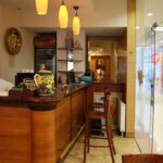 Przytulna kawiarnia Toscana Bistro Bar z drewnianą ladą, lampami wiszącymi i dużym ozdobnym dzbankiem. Wysokie krzesła ustawione wzdłuż lady, a szklane drzwi odsłaniają centrum handlowe. Magazyn z kobietą na okładce spoczywa na małym stoliku na pierwszym planie. projekt i nadzór Tatjana Kobuszewska trksystem.com