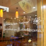 Szklane drzwi z białym odręcznym tekstem w języku polskim promującym stronę na Facebooku, bistrotoscana, obok rysunku kwiatu. W tle Toscana Bistro Bar z przytulnymi drewnianymi meblami, ladą i półkami delikatnie oświetlonymi ciepłym światłem. projekt i nadzór Tatjana Kobuszewska trksystem.com