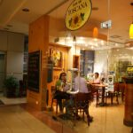 Przeszklona restauracja o nazwie Toscana Bistro Bar przedstawia kilka osób siedzących przy drewnianych stołach, jedzących i rozmawiających. W ciepło oświetlonym wnętrzu znajdują się malowidła ścienne, kredowe menu i rośliny doniczkowe, tworząc przytulną, zachęcającą atmosferę. projekt i nadzór Tatjana Kobuszewska trksystem.com