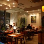 Przytulne, ciepło oświetlone wnętrze baru Toscana Bistro z drewnianymi stołami i krzesłami. Pary siedzą przy oddzielnych stolikach, prowadząc ciche rozmowy. Nowoczesne lampy sufitowe i dekoracje ścienne podkreślają zachęcającą atmosferę, a duże frontowe okna odsłaniają noc na zewnątrz. projekt i nadzór Tatjana Kobuszewska trksystem.com