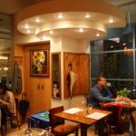 Przytulny, ciepło oświetlony bar Toscana Bistro z dwiema parami jedzącymi przy oddzielnych stolikach. Jedna para rozmawia i trzyma się za ręce, a druga siedzi cicho. Obrazy słoneczników, parasole na ścianach i zakrzywiona oprawa oświetleniowa na suficie tworzą uroczą atmosferę. projekt i nadzór Tatjana Kobuszewska trksystem.com