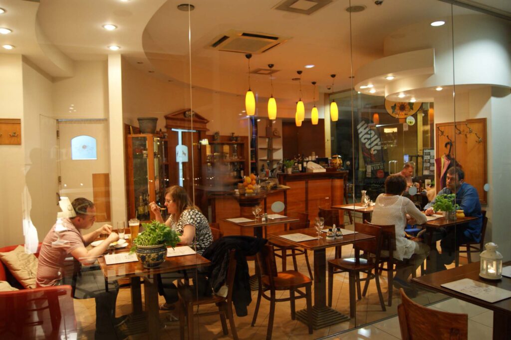 Przytulne wnętrze Toscana Bistro Bar widziane przez szybę, z trzema stolikami zajętymi przez jedzących i rozmawiających ludzi. Ciepłe żółte światła zwisają z sufitu, drewniane meble wypełniają przestrzeń, a rośliny na stołach tworzą zachęcającą, intymną atmosferę. projekt i nadzór Tatjana Kobuszewska trksystem.com