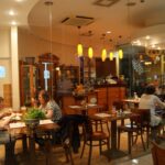 Przytulne wnętrze Toscana Bistro Bar widziane przez szybę, z trzema stolikami zajętymi przez jedzących i rozmawiających ludzi. Ciepłe żółte światła zwisają z sufitu, drewniane meble wypełniają przestrzeń, a rośliny na stołach tworzą zachęcającą, intymną atmosferę. projekt i nadzór Tatjana Kobuszewska trksystem.com