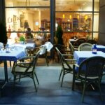 W jadalni na świeżym powietrzu w barze Toscana Bistro znajdują się obrusy w niebieską kratę i wiklinowe krzesła. Stoły oświetlone świecami i białe serwetki tworzą przytulną scenerię, a duże okna oddzielają przytulną przestrzeń wewnętrzną od tętniącej życiem ulicy. projekt i nadzór Tatjana Kobuszewska trksystem.com