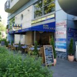 Widok z zewnątrz na Toscana Bistro Bar, restaurację z niebieską markizą i żółtymi wykończeniami, wiklinowymi krzesłami i stolikami na patio, menu na chodniku, nowoczesnym budynkiem, zielenią z przodu i szyldami innych firm. projekt i nadzór Tatjana Kobuszewska trksystem.com