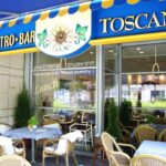 Na zewnątrz baru Toscana Bistro znajdują się wiklinowe krzesła i stoły w niebiesko-białą kratę; duże okna odsłaniają przytulne wnętrze. Powyższy szyld ze słonecznikiem głosi "Toscana". Świeżości dodają bujne zielone rośliny. projekt i nadzór Tatjana Kobuszewska trksystem.com