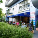 Ludzie siedzą przy stolikach na świeżym powietrzu pod niebieskimi markizami w Toscana Bistro Bar, mieszczącym się w nowoczesnym budynku z różnymi szyldami w języku polskim. Patio otoczone jest zielonymi krzewami, a przy wejściu znajduje się tablica z menu. Drzewa i zieleń widoczne są po lewej stronie. projekt i nadzór Tatjana Kobuszewska trksystem.com