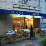 Na zewnątrz Toscana Bistro Bar znajdują się wiklinowe krzesła i stoliki w niebieską kratę. Niebieska markiza ocienia obszar, z roślinami doniczkowymi i ciepło oświetlonym wnętrzem widocznym przez okna. Powyżej znajduje się studio masażu tajskiego z widokiem na scenę. projekt i nadzór Tatjana Kobuszewska trksystem.com