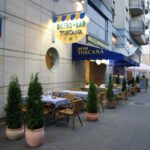 Bar Toscana Bistro oferuje miejsca do siedzenia na świeżym powietrzu na miejskim chodniku, z niebieskimi obrusami i wiklinowymi krzesłami wśród zieleni doniczkowej. Szyld bistro świeci nad markizą w niebieskie paski, otoczoną nowoczesnymi mieszkaniami i znakiem ulicznym. projekt i nadzór Tatjana Kobuszewska trksystem.com