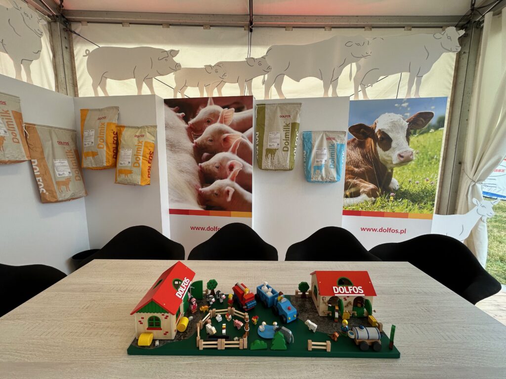 Na stole z jasnego drewna w stoisku targowym Szepietowo stoi diorama farmy z figurkami zwierząt i traktorów. Za nią plakaty przedstawiające świnie i cielaka z workami paszy i papierowymi wycinkami zwierząt wzdłuż tylnej ściany namiotu. Na szyldach widnieje napis "Dolfos". projekt i nadzór Tatjana Kobuszewska