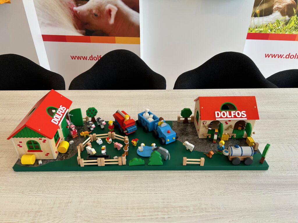 Diorama farmy z dwoma budynkami z czerwonym dachem z napisem DOLFOS, zabawkowymi zwierzętami, pojazdami, drzewami i płotami znajduje się na zielonej podstawie. To stoisko reklamowe Dolfos było prezentowane na Regionalnej Wystawie Zwierząt Hodowlanych. projekt i nadzór Tatjana Kobuszewska