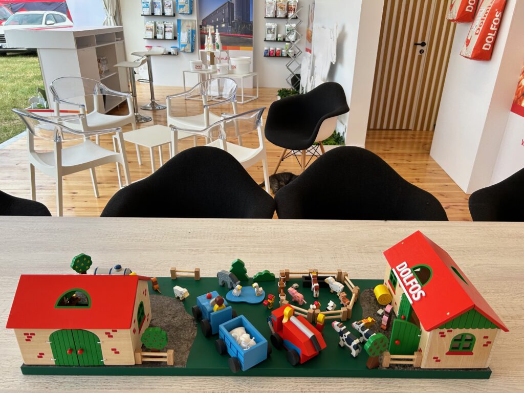 Miniaturowy model farmy ze stodołami z napisem DOLFOS znajduje się na szczycie stołu. W tle stoisko reklamowe Dolfos z białymi krzesłami, broszurami i drewnianą podłogą, łączące zabawny urok z profesjonalną prezentacją. projekt i nadzór Tatjana Kobuszewska